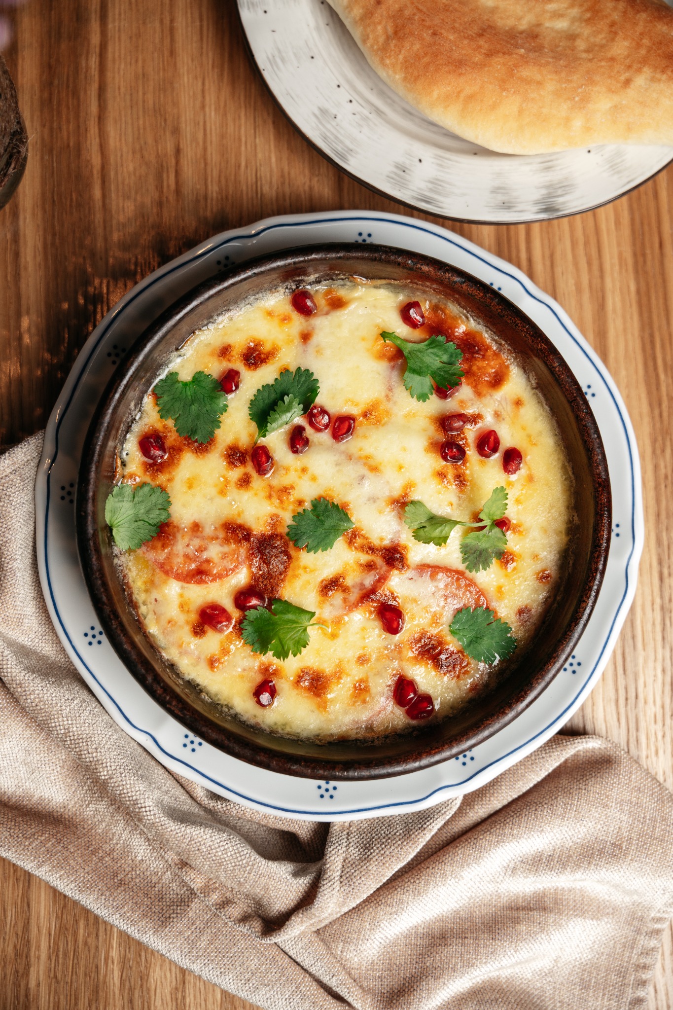 Kremowe danie w glinianej miseczce, udekorowane granatem i kolendrą, na drewnianym stole z chlebem i lnianą ściereczką.