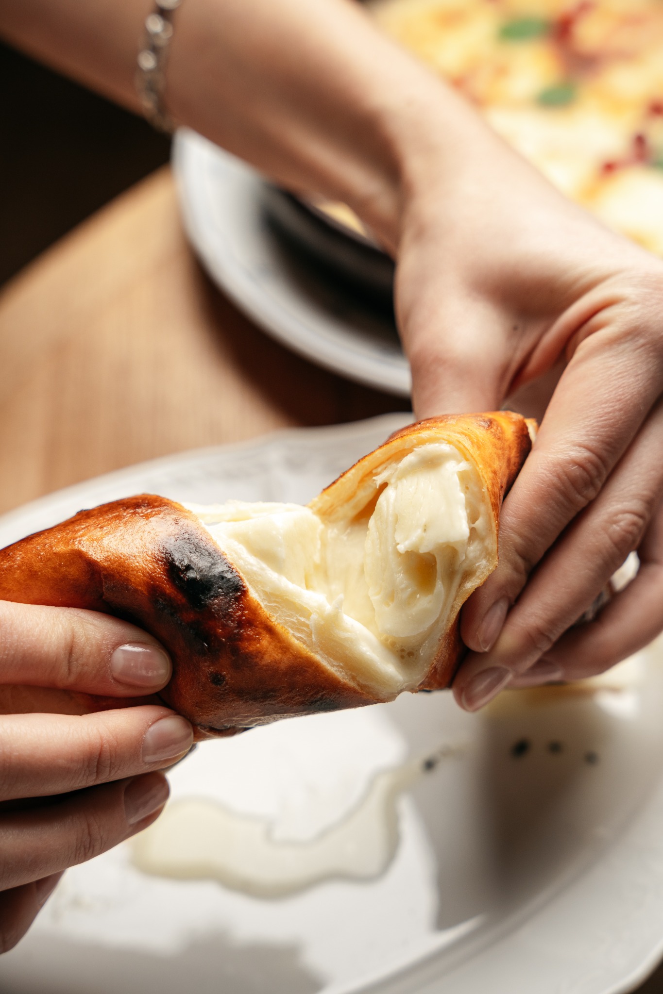Dwie dłonie rozrywają na pół chrupiące ciasto wypełnione kremowym serem, z widocznym nadzieniem i topiącym się serem na talerzu.