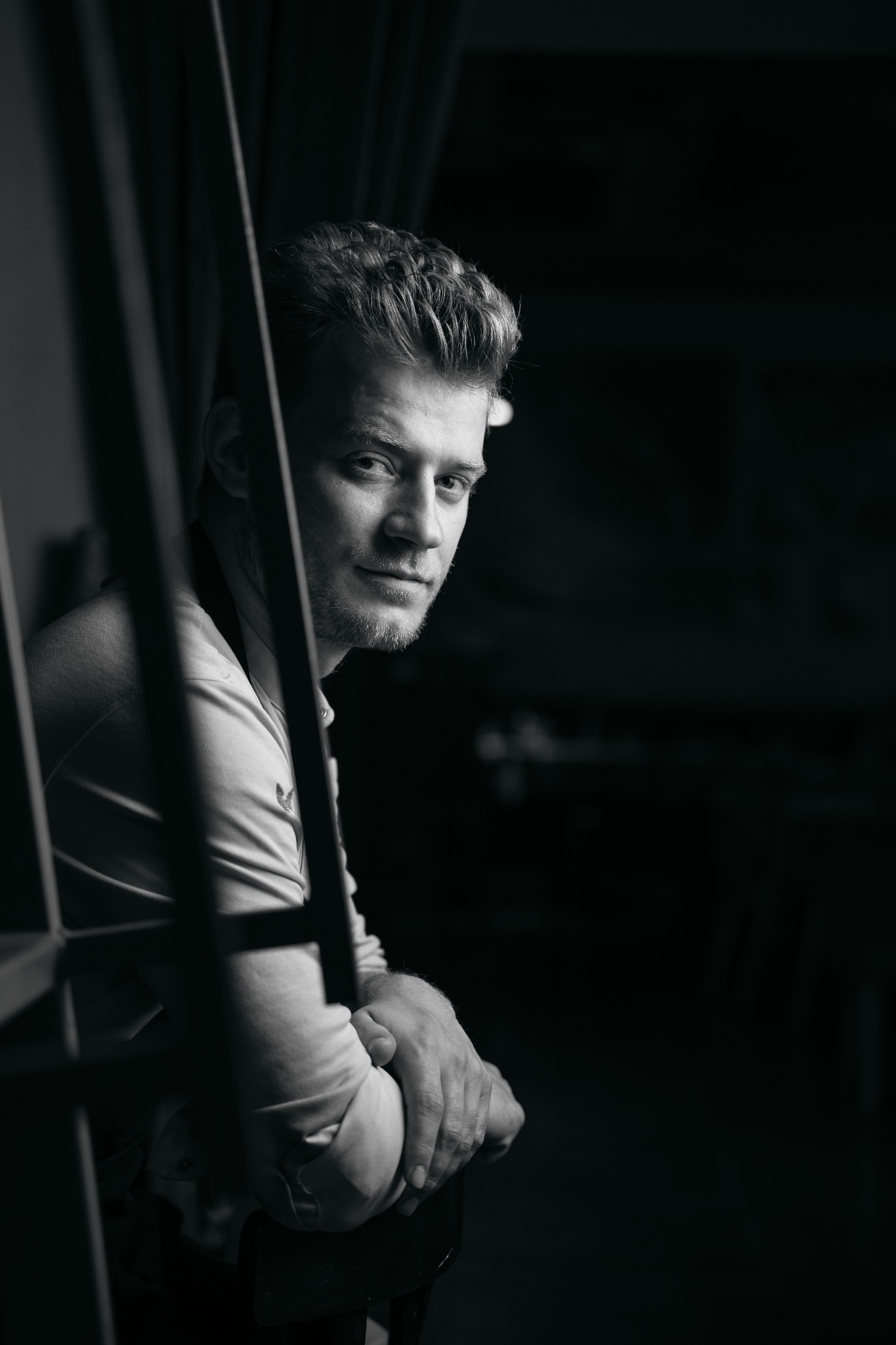 Portret mężczyzny w czerni i bieli, lekko zamyślony, z dłońmi splecionymi na oparciu krzesła, widziany przez metalową konstrukcję, z ciemnym tłem.