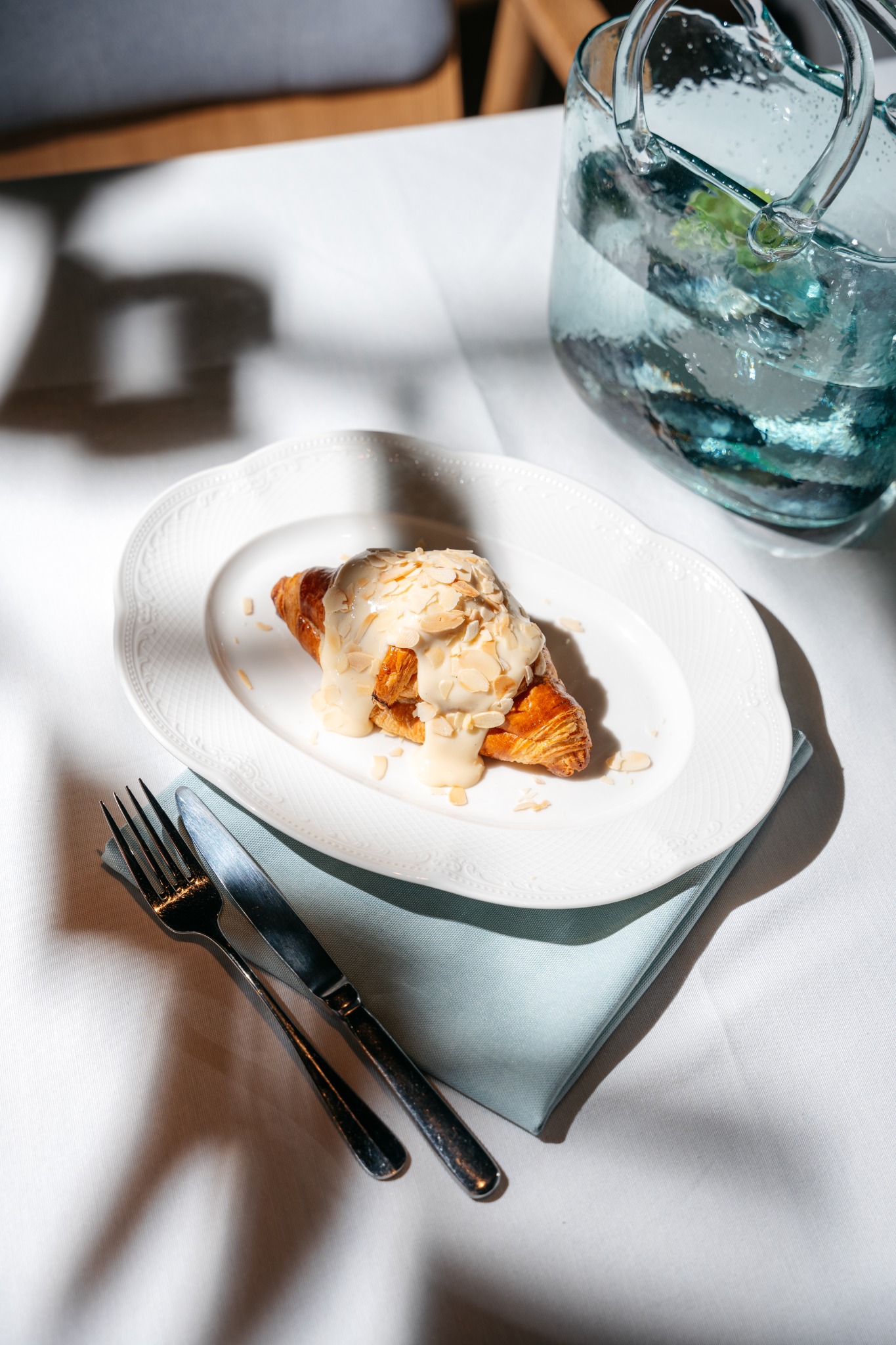 Kremowy rogalik z płatkami migdałów na białym talerzu, obok sztućce i dzbanek z wodą, na tle białego obrusu i jasnoniebieskiej serwetki.