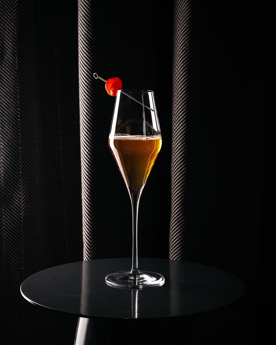 Elegancki kieliszek z bursztynowym napojem i dekoracją z czerwonej wiśni na szpilce, na czarnym okrągłym stoliku z ciemnym, wzorzystym tłem.