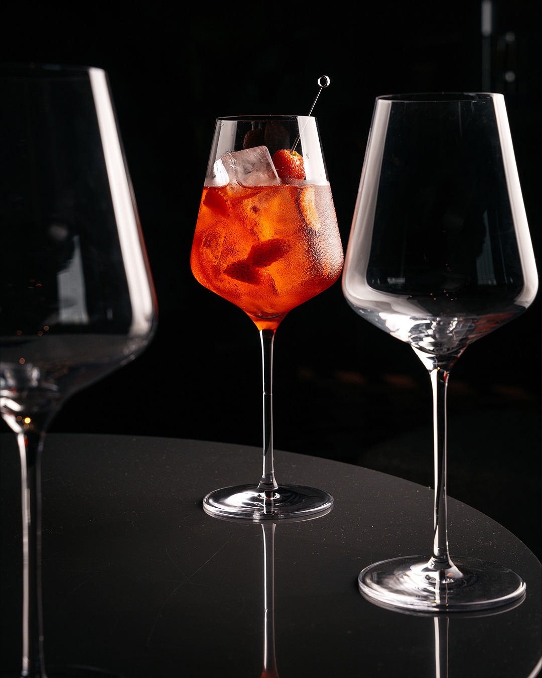 Oranżowy koktajl Aperol Spritz z kostkami lodu i truskawką na szpilce w szklance o gruszkowatym kształcie, otoczony dwiema pustymi szklankami na wysokich nóżkach, ustawiony na błyszczącym czarnym...