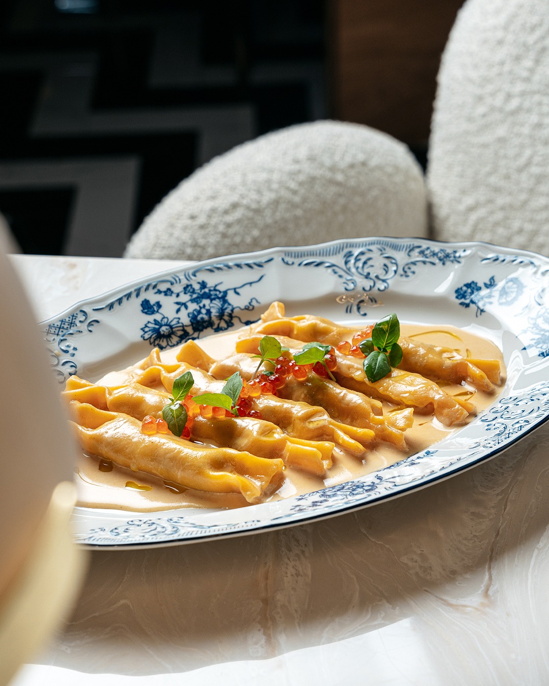 Elegancko podane ravioli z kawiorem z łososia i listkami bazylii na ozdobnym talerzu z motywem kwiatowym, ustawione na marmurowym blacie.