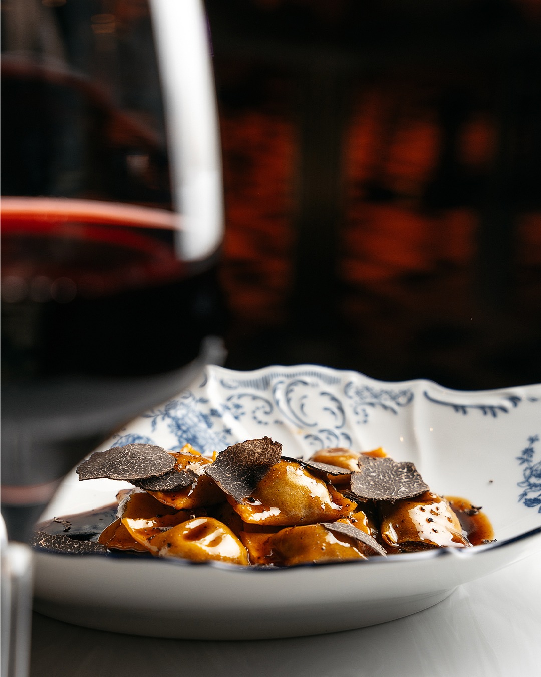 Eleganckie ravioli z truflami na dekoracyjnym talerzu, z kieliszkiem czerwonego wina w tle.