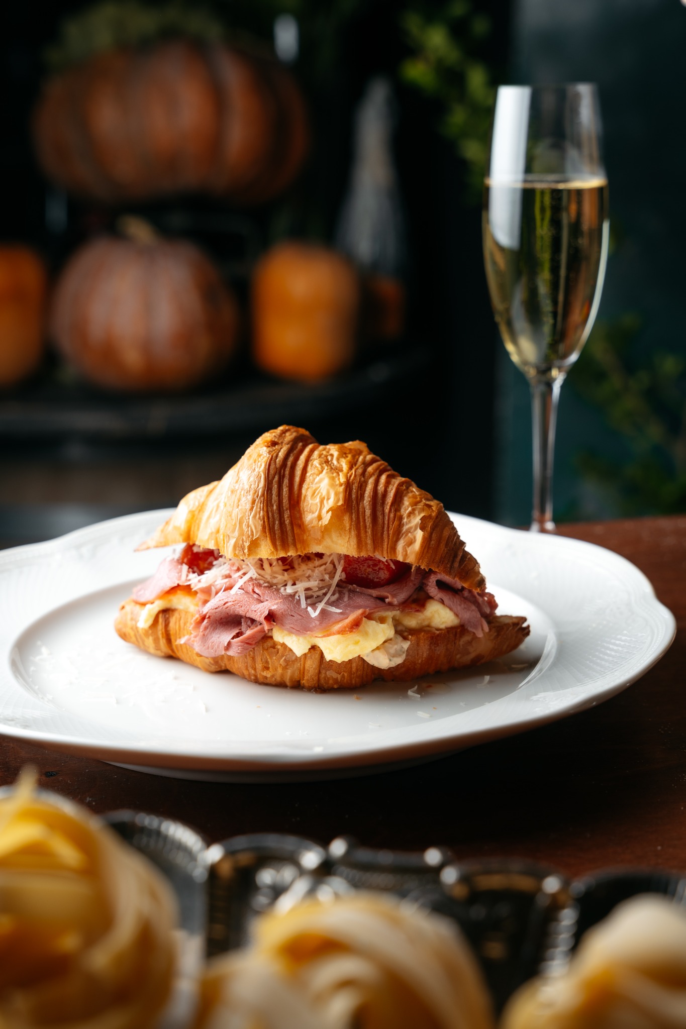 Kulinarna martwa natura: Croissant z szynką, serem i warzywami na białym talerzu, w tle kieliszek szampana i dynie.