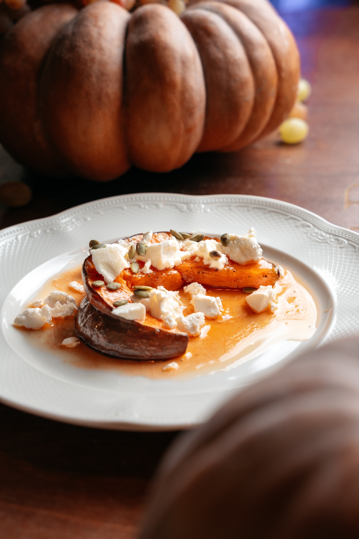 Dwa plastry pieczonej dyni na białym talerzu, polane sosem i posypane serem feta oraz pestkami dyni, w tle dynia i winogrona na drewnianym stole.