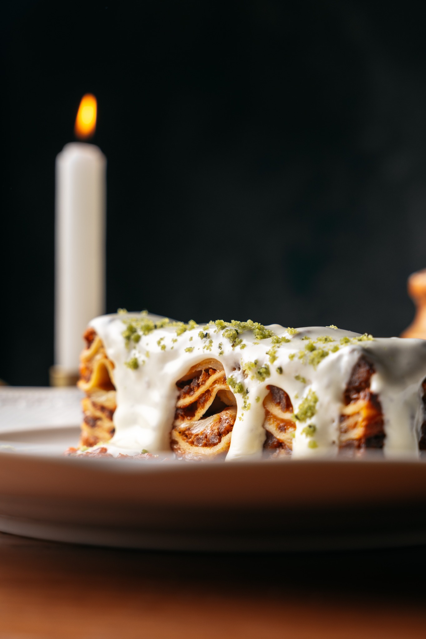 Apetyczna lasagne z kremowym sosem i posypką pistacjową na talerzu, w tle płonąca świeca.