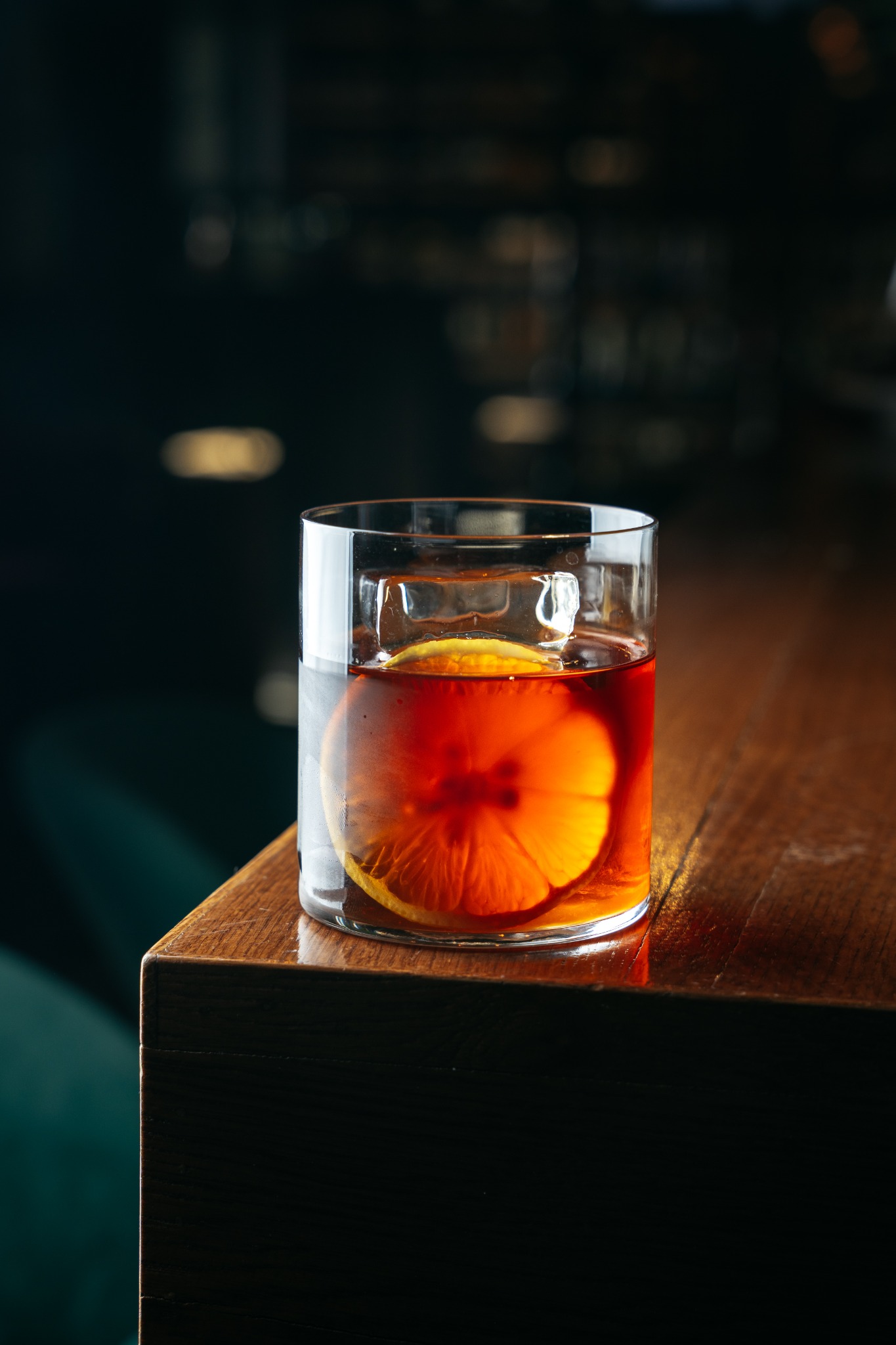 Kieliszek z bursztynowym płynem, kostką lodu i plasterkiem pomarańczy na drewnianym blacie baru, w tle rozmyte światła.