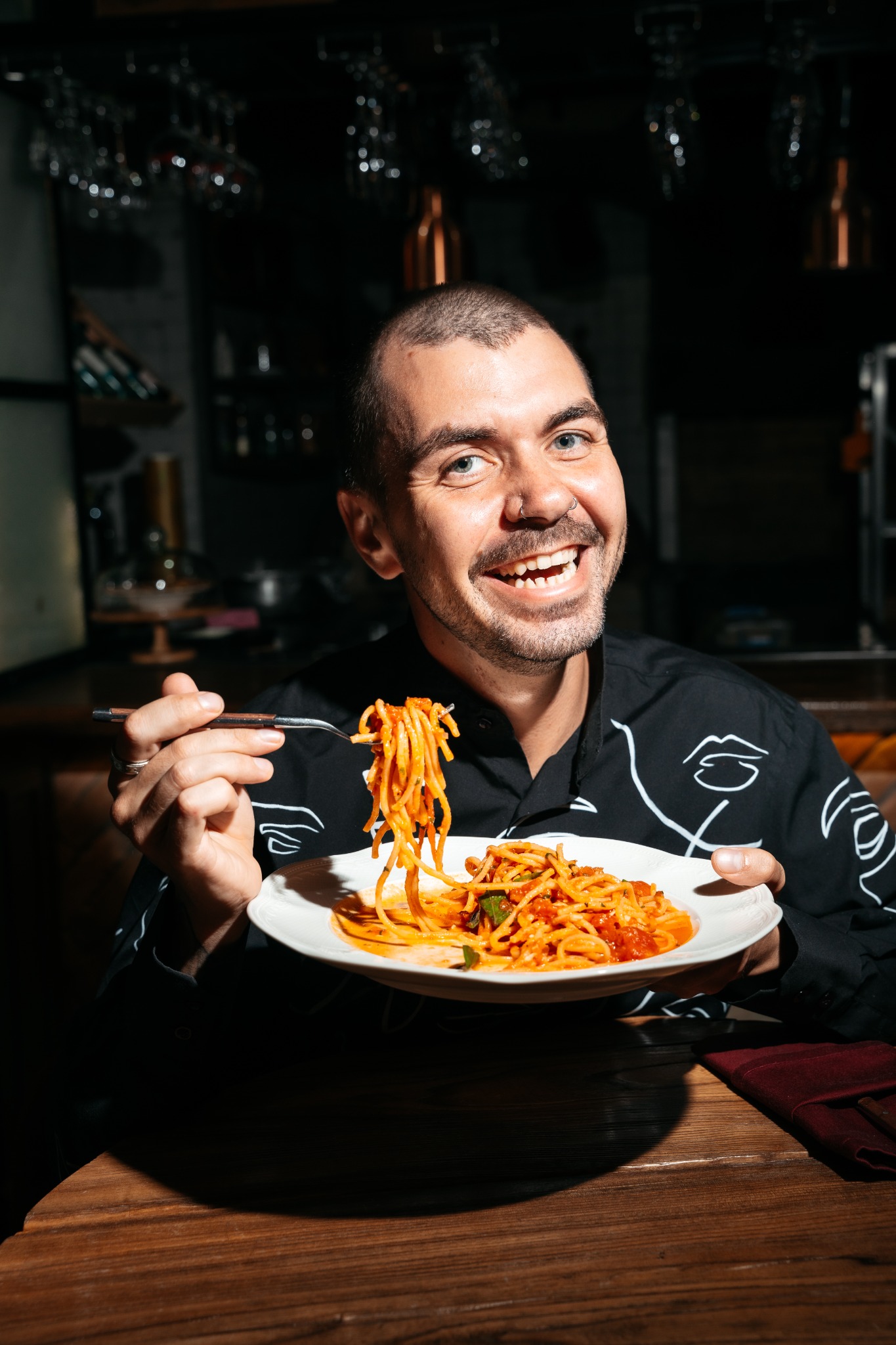Uśmiechnięty mężczyzna z kolczykiem w nosie, w czarnej koszuli z białymi wzorami, trzyma widelec z makaronem spaghetti nad talerzem pełnym spaghetti z sosem pomidorowym w restauracji.