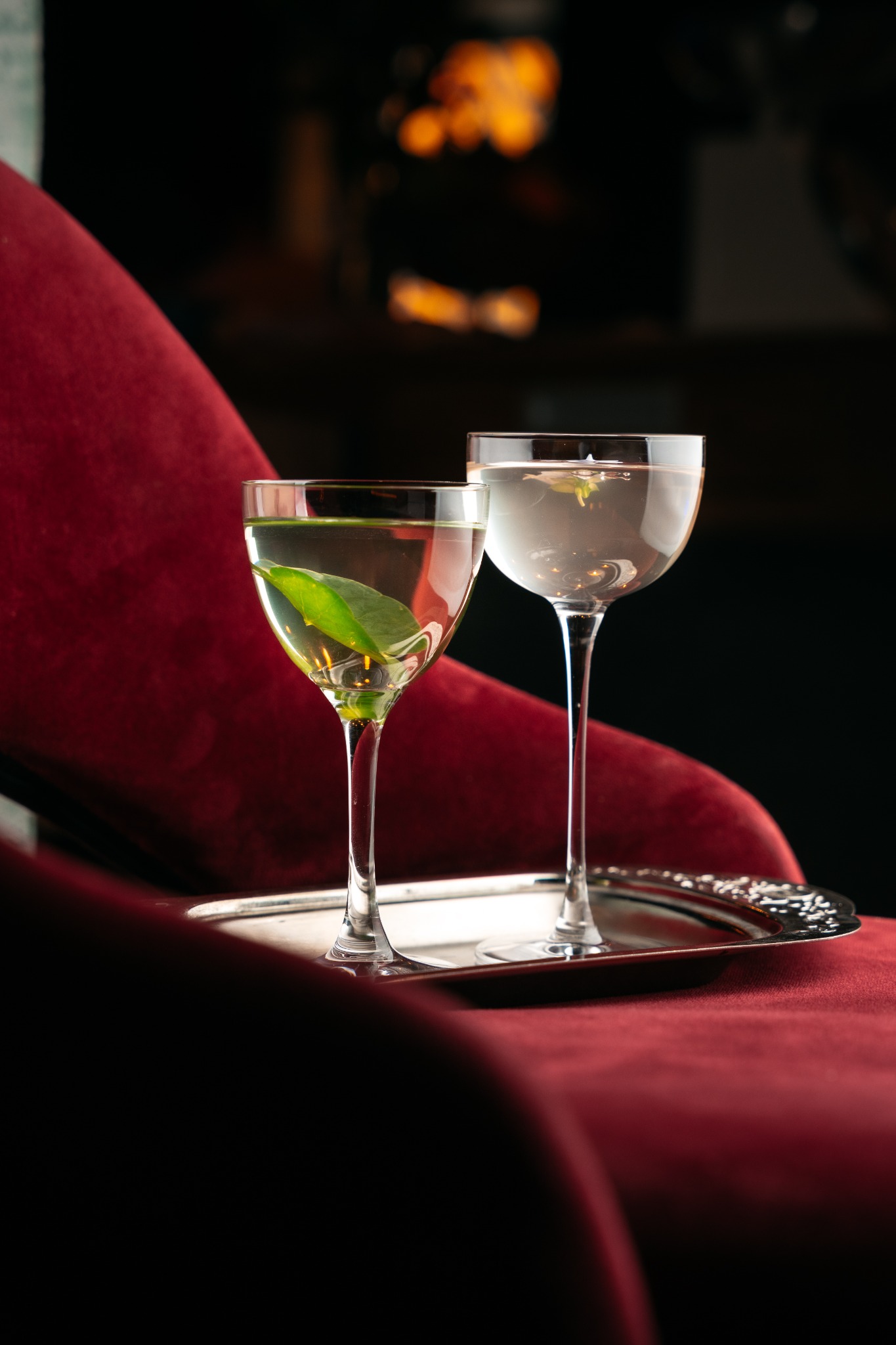 Dwa koktajle w kieliszkach typu coupe, jeden z liściem, drugi z kwiatem, podane na srebrnej tacy na tle czerwonego, aksamitnego fotela.