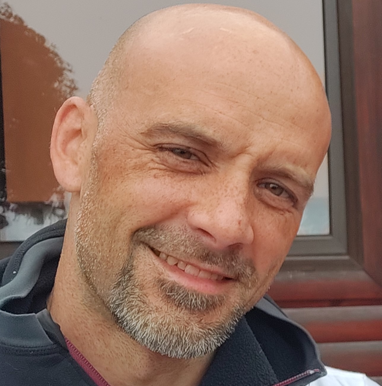 Portret uśmiechniętego łysiejącego mężczyzny z siwiejącą brodą, na tle brązowej ściany i okna.