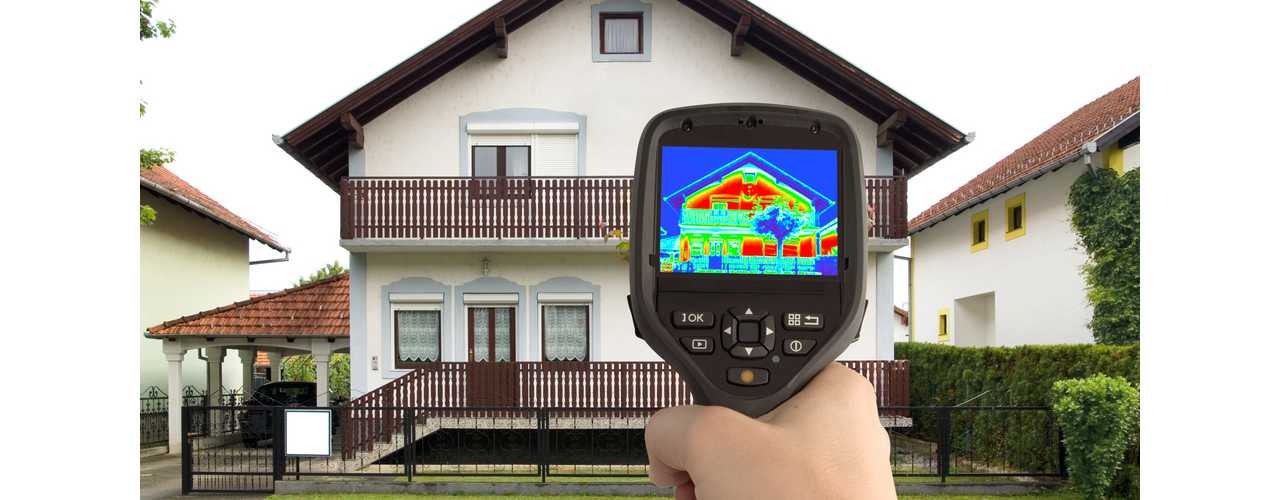 Termowizja domu jednorodzinnego z balkonem, widoczna na ekranie kamery termowizyjnej trzymanej w dłoni.