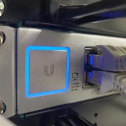 Szczegółowe ujęcie portu sieciowego z podłączonymi kablami Ethernet, diody sygnalizujące status połączenia, obudowa urządzenia z logo podświetlonym na niebiesko.