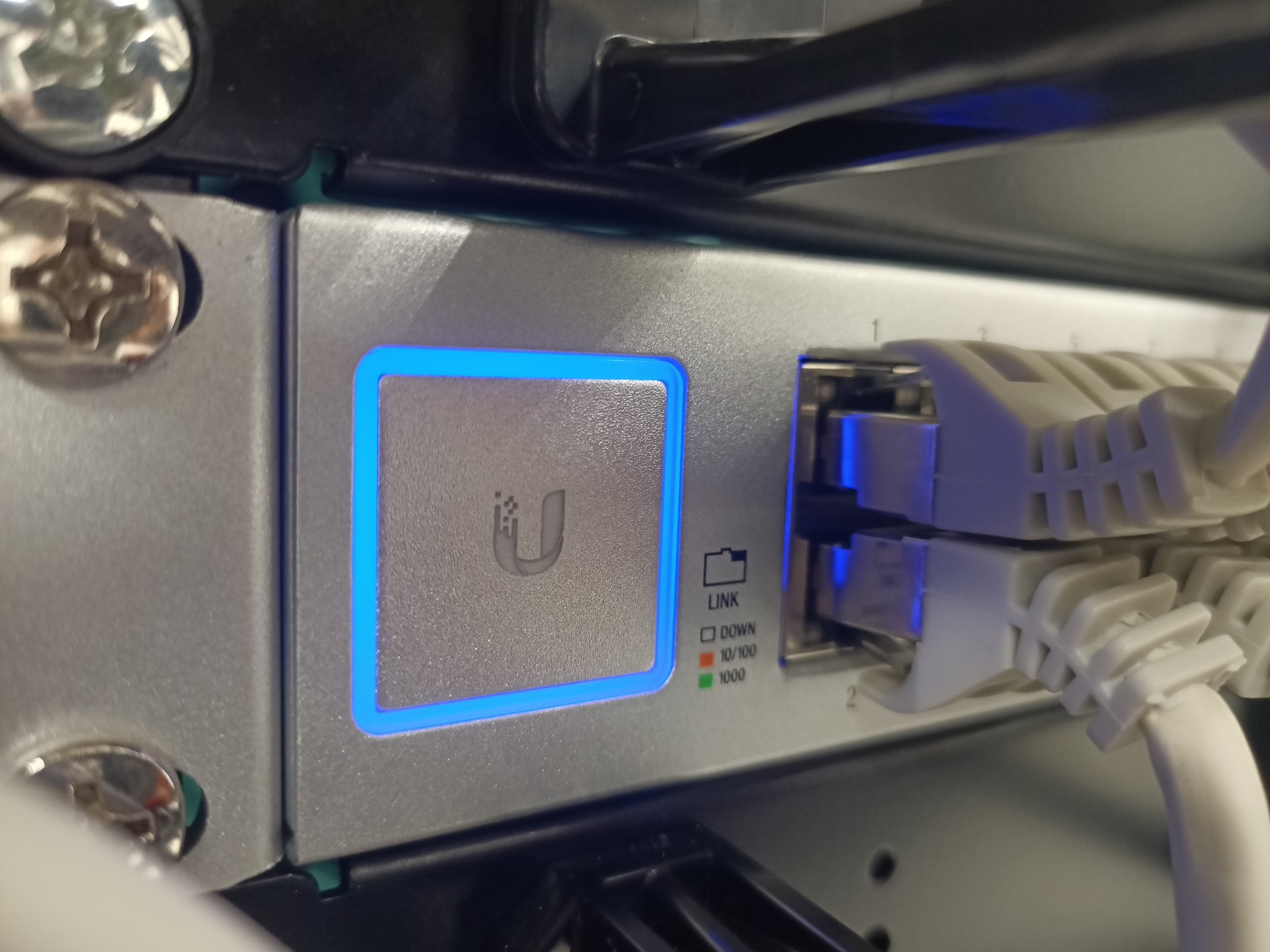 Szczegółowe ujęcie portu sieciowego z podłączonymi kablami Ethernet, diody sygnalizujące status połączenia, obudowa urządzenia z logo podświetlonym na niebiesko.