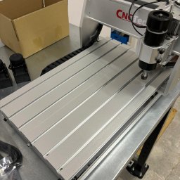 AD COMO GABRIELA SZEREMETA - Srebrna frezarka CNC z czarnym wrzecionem, widoczna na metalowym stole, obok kartonowe pudełko i akcesoria. Maszyna z napisem CNC-Tec.