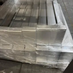 Stos aluminiowych obróbek blacharskich, zabezpieczonych folią, gotowych do transportu. Widoczne łączenia elementów i surowy wygląd metalu.