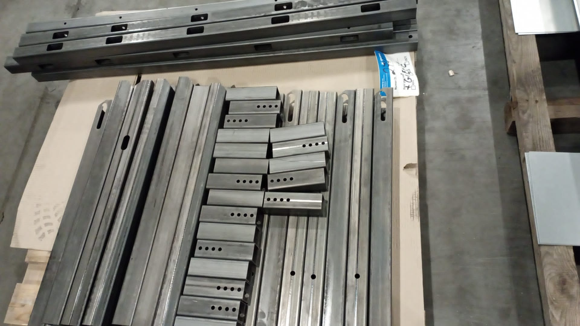 Metalowe profile i elementy konstrukcyjne z otworami, ułożone na palecie w hali produkcyjnej, gotowe do dalszej obróbki lub montażu. Widoczne detale precyzyjnego wykonania.