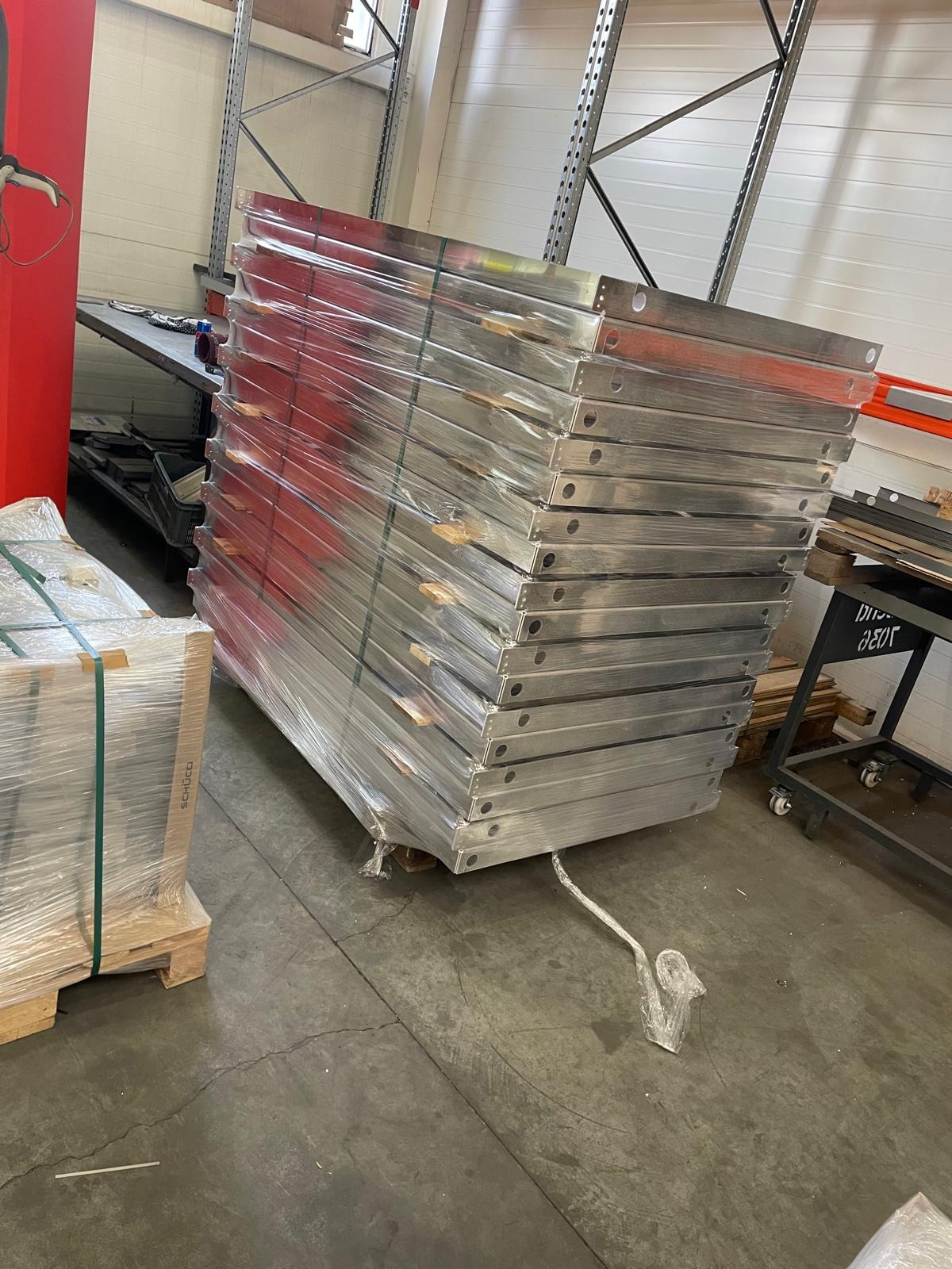 Stos aluminiowych profili, prawdopodobnie parapetów, owiniętych folią stretch, składowanych w magazynie. Widoczne otwory montażowe.