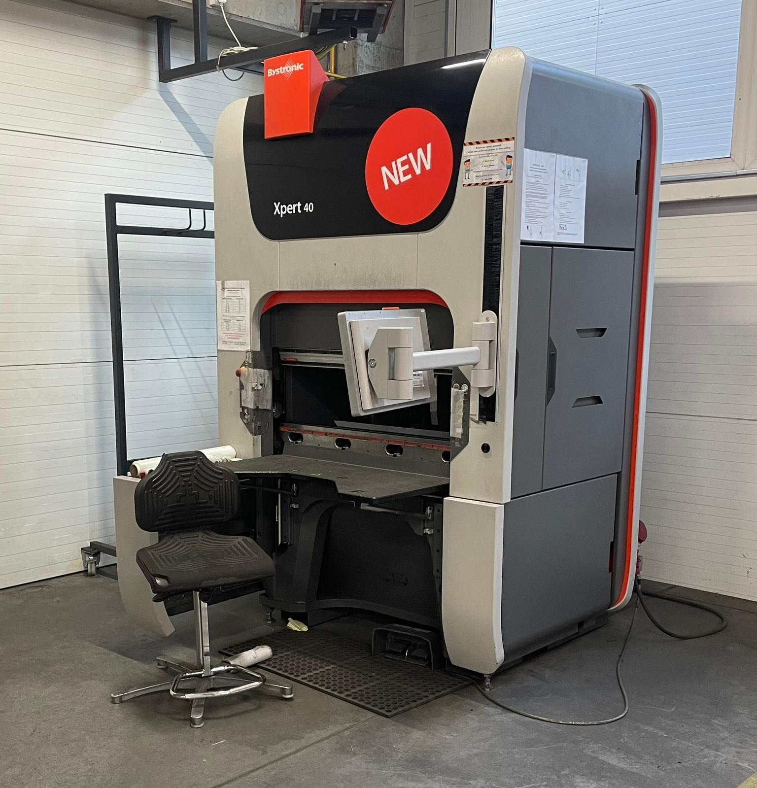 Nowoczesna prasa krawędziowa Bystronic Xpert 40 z pomarańczowym logo i naklejką 'NEW', widoczna w hali produkcyjnej z szarą podłogą i białymi ścianami.