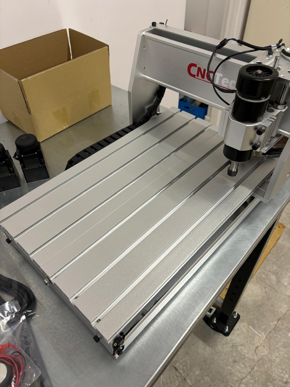 Srebrna frezarka CNC z czarnym wrzecionem, widoczna na metalowym stole, obok kartonowe pudełko i akcesoria. Maszyna z napisem CNC-Tec.