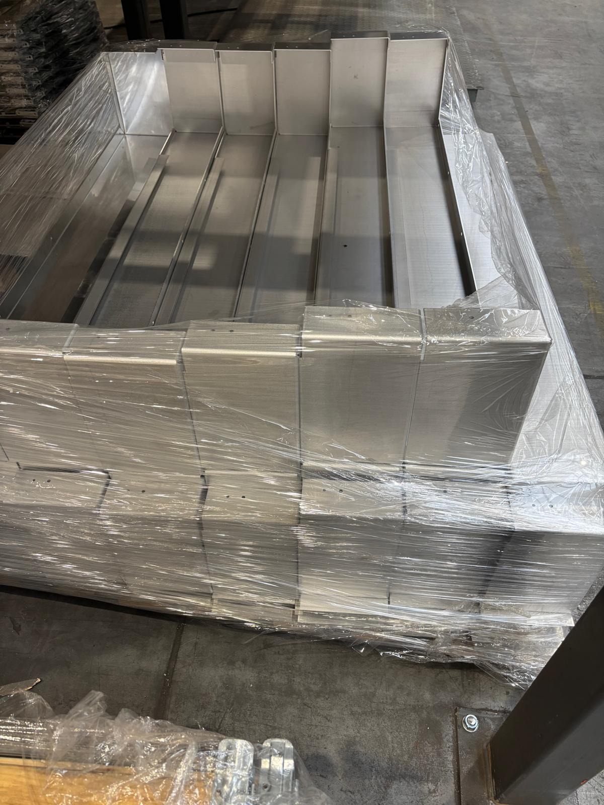 Stos aluminiowych obróbek blacharskich, zabezpieczonych folią, gotowych do transportu. Widoczne łączenia elementów i surowy wygląd metalu.