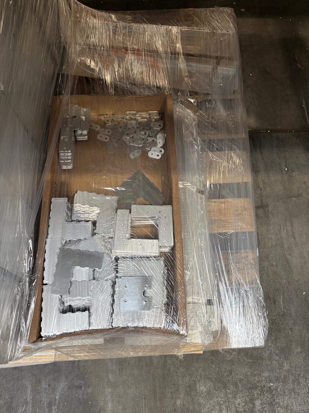 Paleta z metalowymi elementami konstrukcyjnymi, zapakowana w folię. Widoczne różnorodne kształty i rozmiary, niektóre z otworami, sugerujące elementy montażowe.