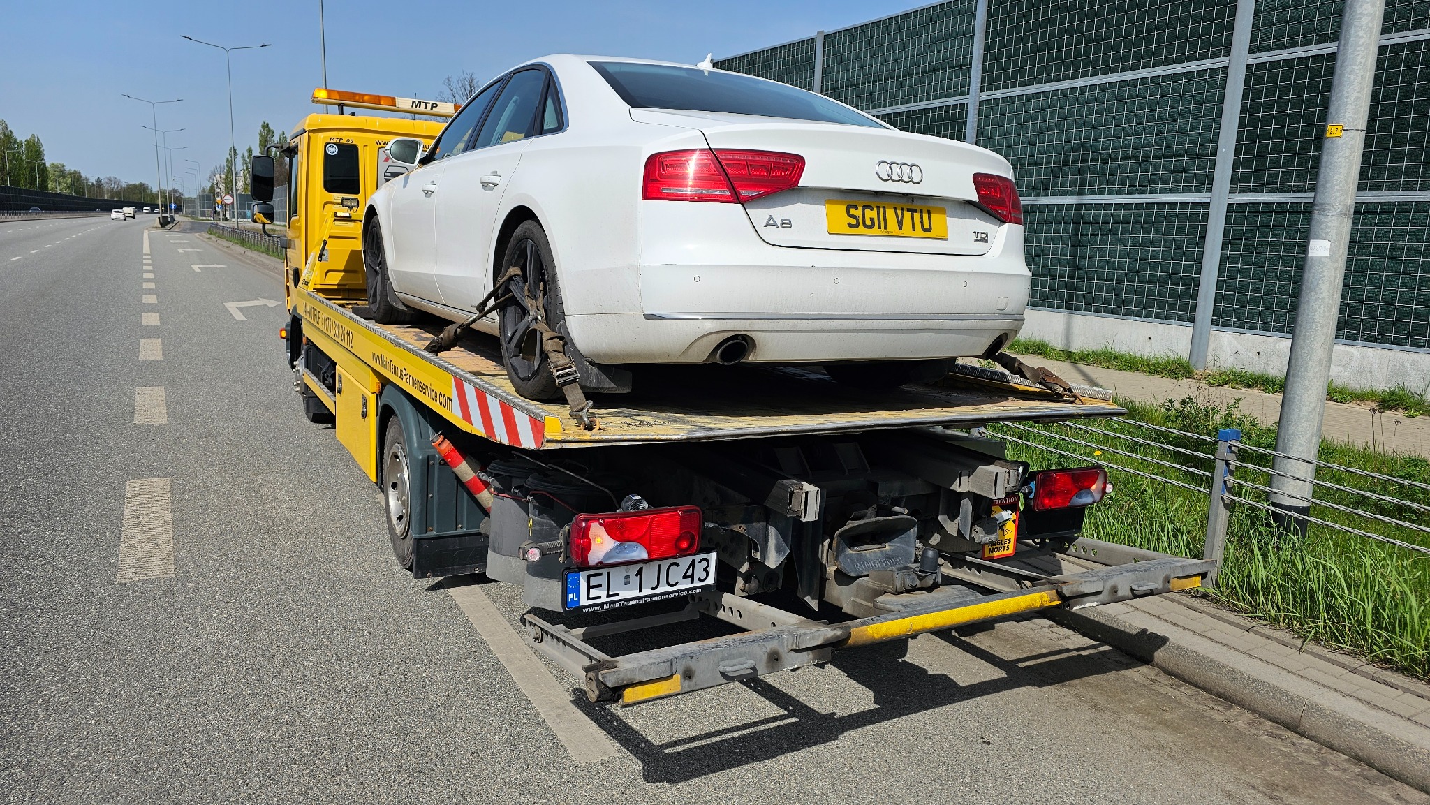Żółta laweta z białym Audi A8 na platformie, zabezpieczonym pasami transportowymi, na poboczu drogi z ekranami akustycznymi w tle.