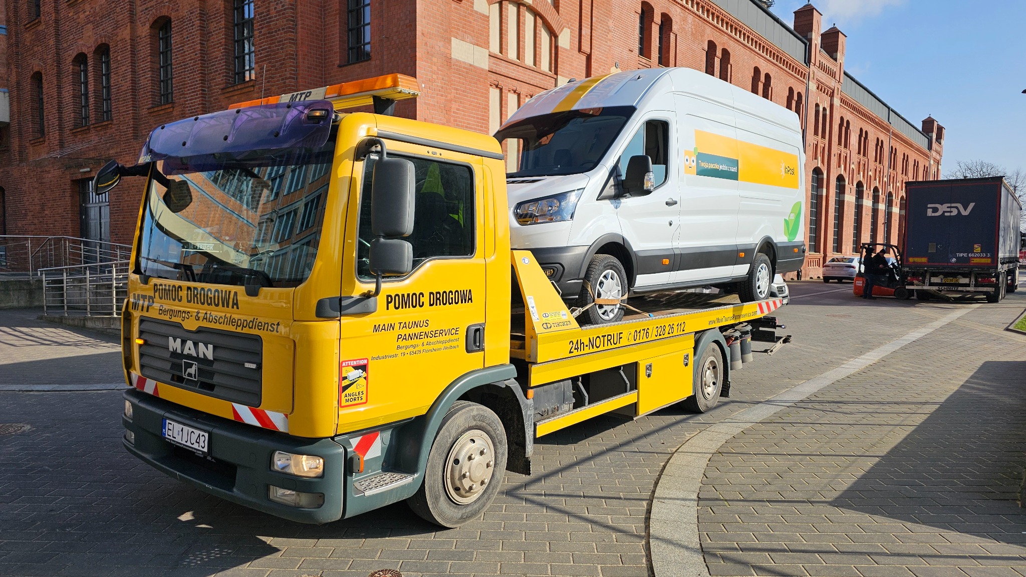Żółta laweta MAN z napisem POMOC DROGOWA przewozi białego busa InPost na tle ceglanego budynku.