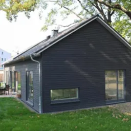 Nowoczesny, parterowy dom z ciemną, drewnianą elewacją, widoczny z boku na tle zielonego trawnika i drzew. Na tarasie zestaw mebli ogrodowych.