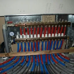 Rozdzielacz ogrzewania podłogowego z licznymi rurami w kolorach czerwonym i niebieskim, zamontowany w metalowej obudowie w surowym stanie wykończenia wnętrza.