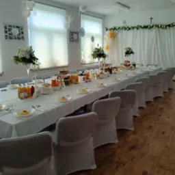 Elegancko nakryty stół na uroczystość: biały obrus, kwiaty, napoje, dania na talerzach. Krzesła z pokrowcami, dekoracje z balonów i zieleni w tle.