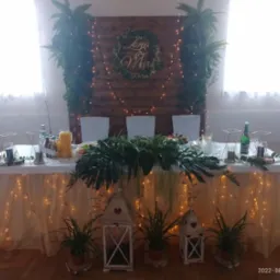 Dekoracja stołu weselnego z drewnianym tłem, roślinami, lampionami i delikatnym oświetleniem, tworzącą elegancką i naturalną atmosferę.