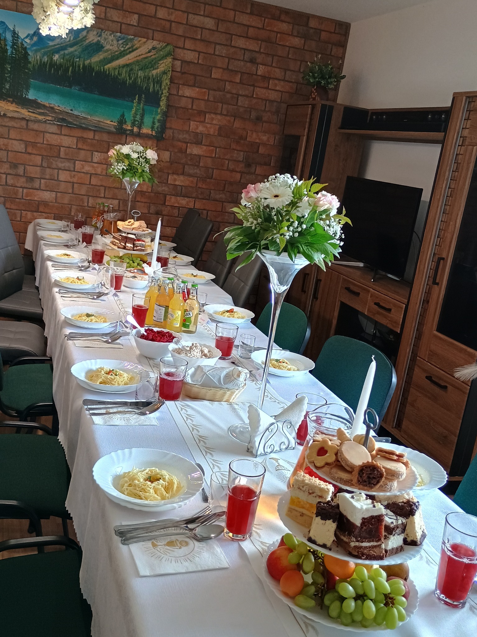 Uroczysty stół z nakryciami, dekoracjami kwiatowymi, ciastami i owocami, przygotowany do przyjęcia gości. W tle ceglana ściana i meble.