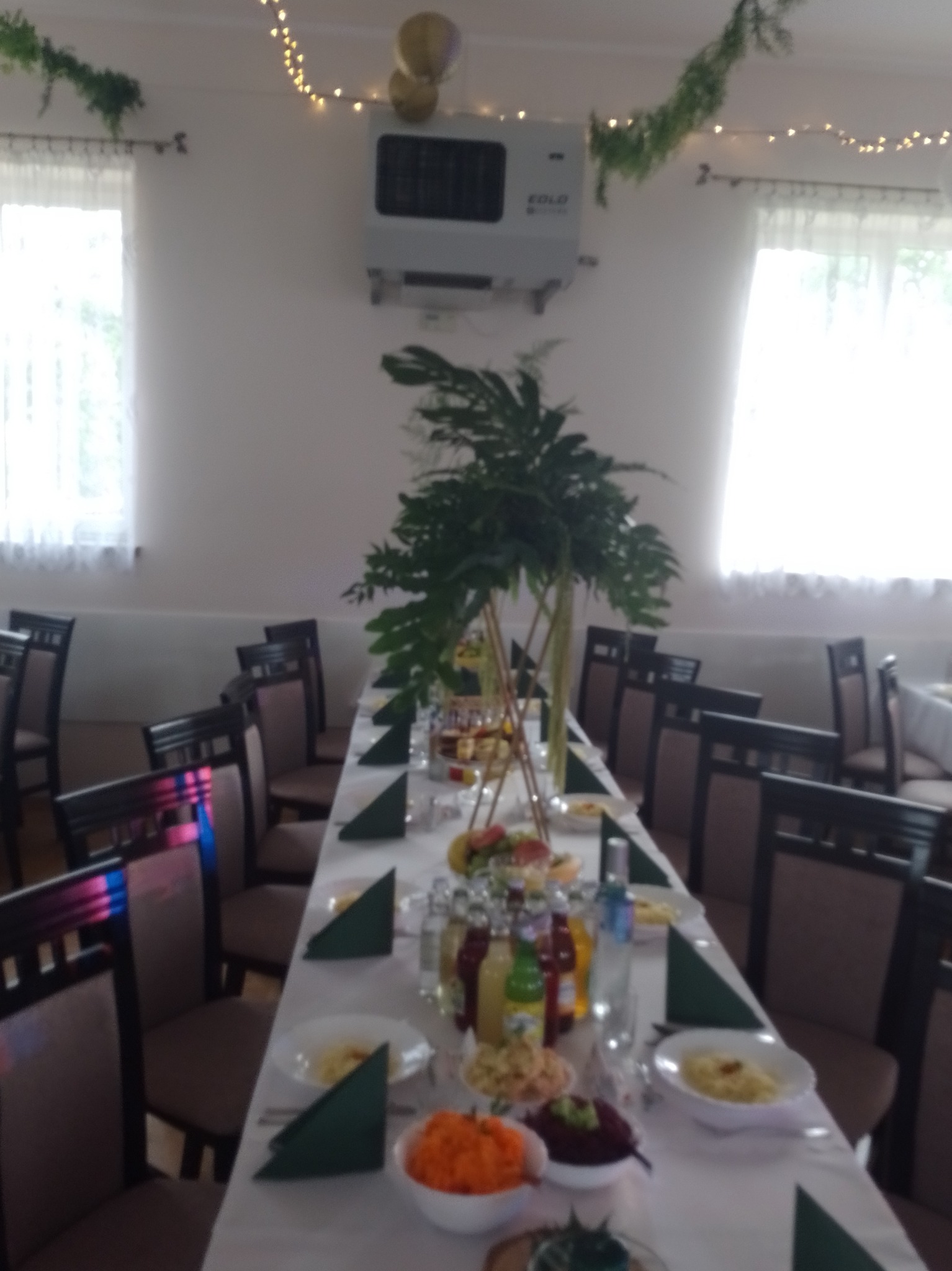 Sala bankietowa z długim stołem, nakryta białym obrusem i zielonymi serwetkami, udekorowana kwiatami i girlandami świetlnymi, gotowa na uroczystość.