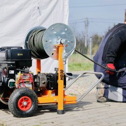 EDKAN SPÓŁKA Z OGRANICZONĄ ODPOWIEDZIALNOŚCIĄ - Hydraulik w kombinezonie roboczym obsługuje mobilną maszynę do czyszczenia rur z nawiniętym wężem, na zewnątrz, w tle biały namiot i samochód.