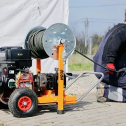 Hydraulik w kombinezonie roboczym obsługuje mobilną maszynę do czyszczenia rur z nawiniętym wężem, na zewnątrz, w tle biały namiot i samochód.