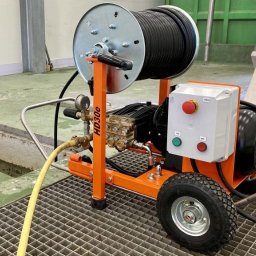 EDKAN SPÓŁKA Z OGRANICZONĄ ODPOWIEDZIALNOŚCIĄ - Pomarańczowa maszyna HD30e do udrażniania rur z nawiniętym wężem na szpuli, stojąca na metalowej kracie obok otwartego włazu kanalizacyjnego.