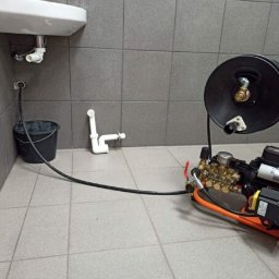 EDKAN SPÓŁKA Z OGRANICZONĄ ODPOWIEDZIALNOŚCIĄ - Profesjonalna maszyna wysokociśnieniowa do czyszczenia rur podłączona do odpływu umywalki, z wiaderkiem na ścieki i fragmentem rury na podłodze w łazience z szarymi płytkami.