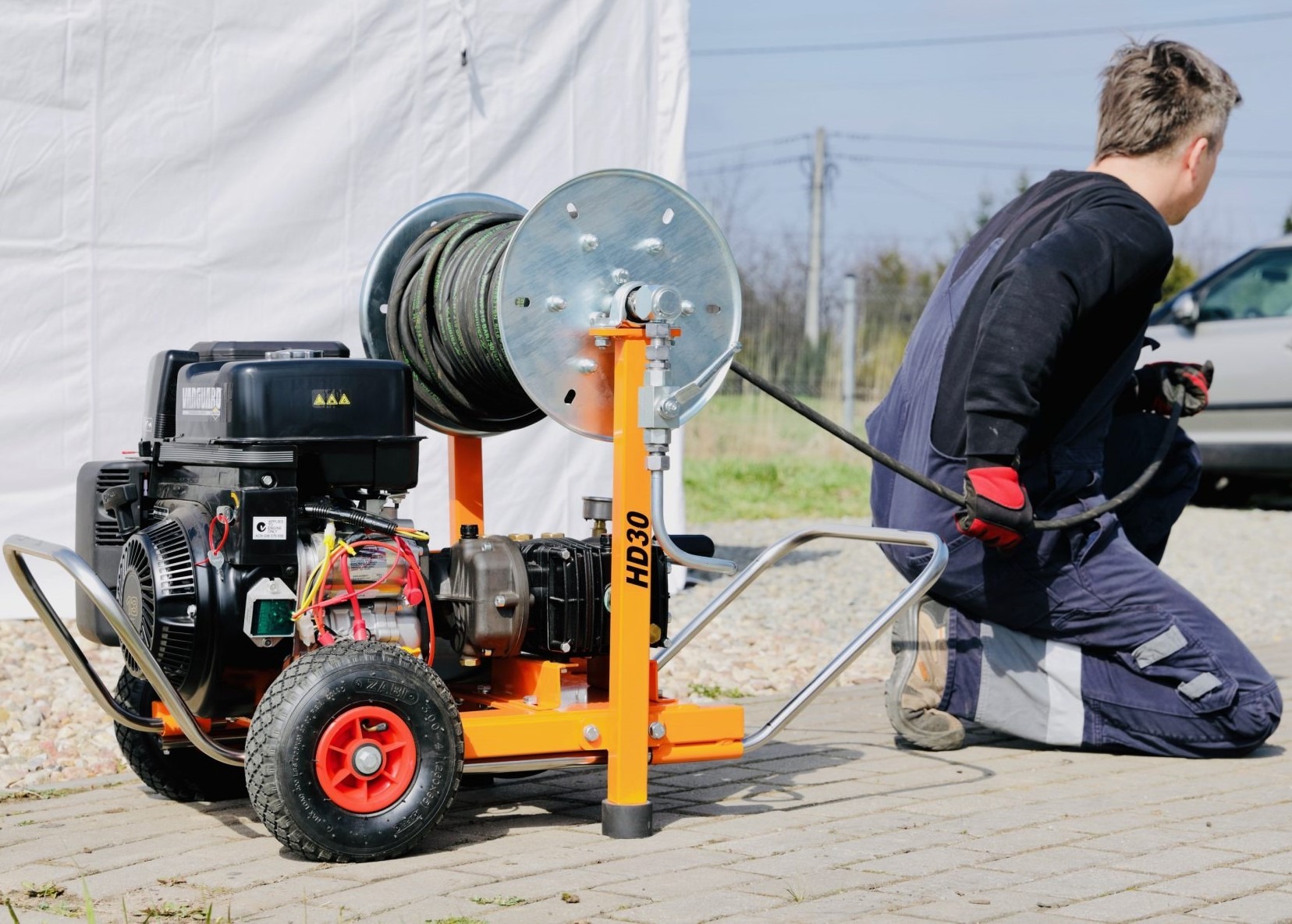 Hydraulik w kombinezonie roboczym obsługuje mobilną maszynę do czyszczenia rur z nawiniętym wężem, na zewnątrz, w tle biały namiot i samochód.