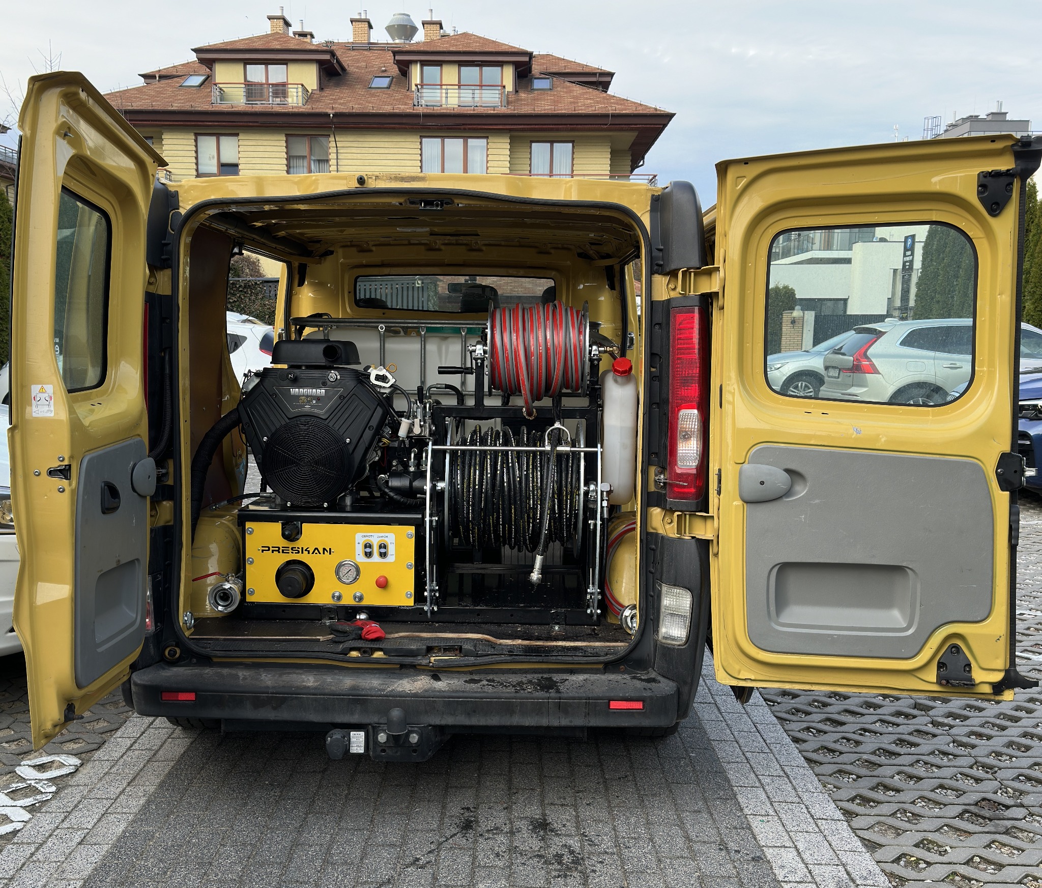 Żółty samochód dostawczy z otwartymi tylnymi drzwiami, wewnątrz widoczny agregat wysokociśnieniowy z bębnami na węże, na zewnątrz budynek mieszkalny i zaparkowane samochody.