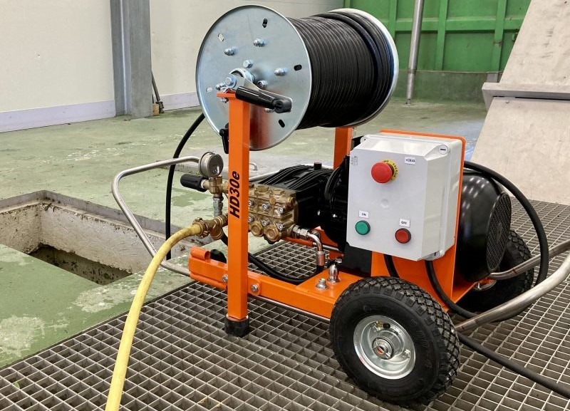 Pomarańczowa maszyna HD30e do udrażniania rur z nawiniętym wężem na szpuli, stojąca na metalowej kracie obok otwartego włazu kanalizacyjnego.