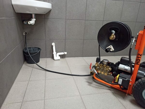 Profesjonalna maszyna wysokociśnieniowa do czyszczenia rur podłączona do odpływu umywalki, z wiaderkiem na ścieki i fragmentem rury na podłodze w łazience z szarymi płytkami.
