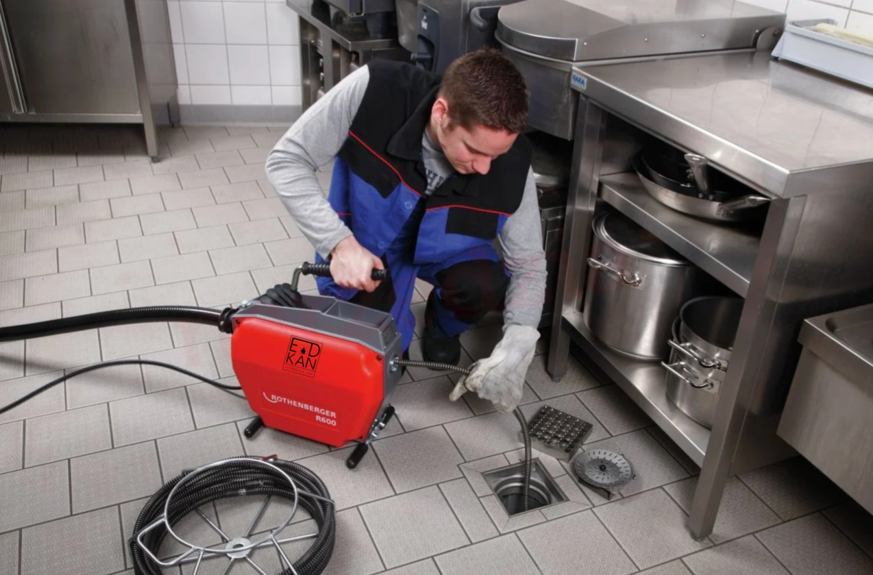 Hydraulik w rękawicach roboczych udrażnia odpływ podłogowy w kuchni przemysłowej za pomocą elektrycznej sprężyny Rothenberger R600.