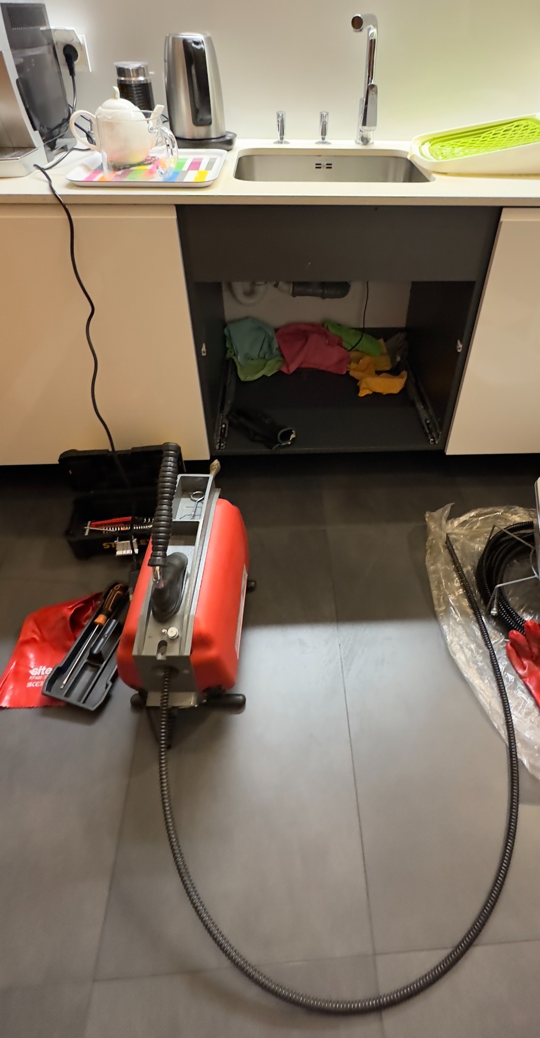 Sprzęt do czyszczenia rur kanalizacyjnych, z widoczną sprężyną wprowadzana do syfonu pod zlewem, obok skrzynka z narzędziami i komplet uszczelek.