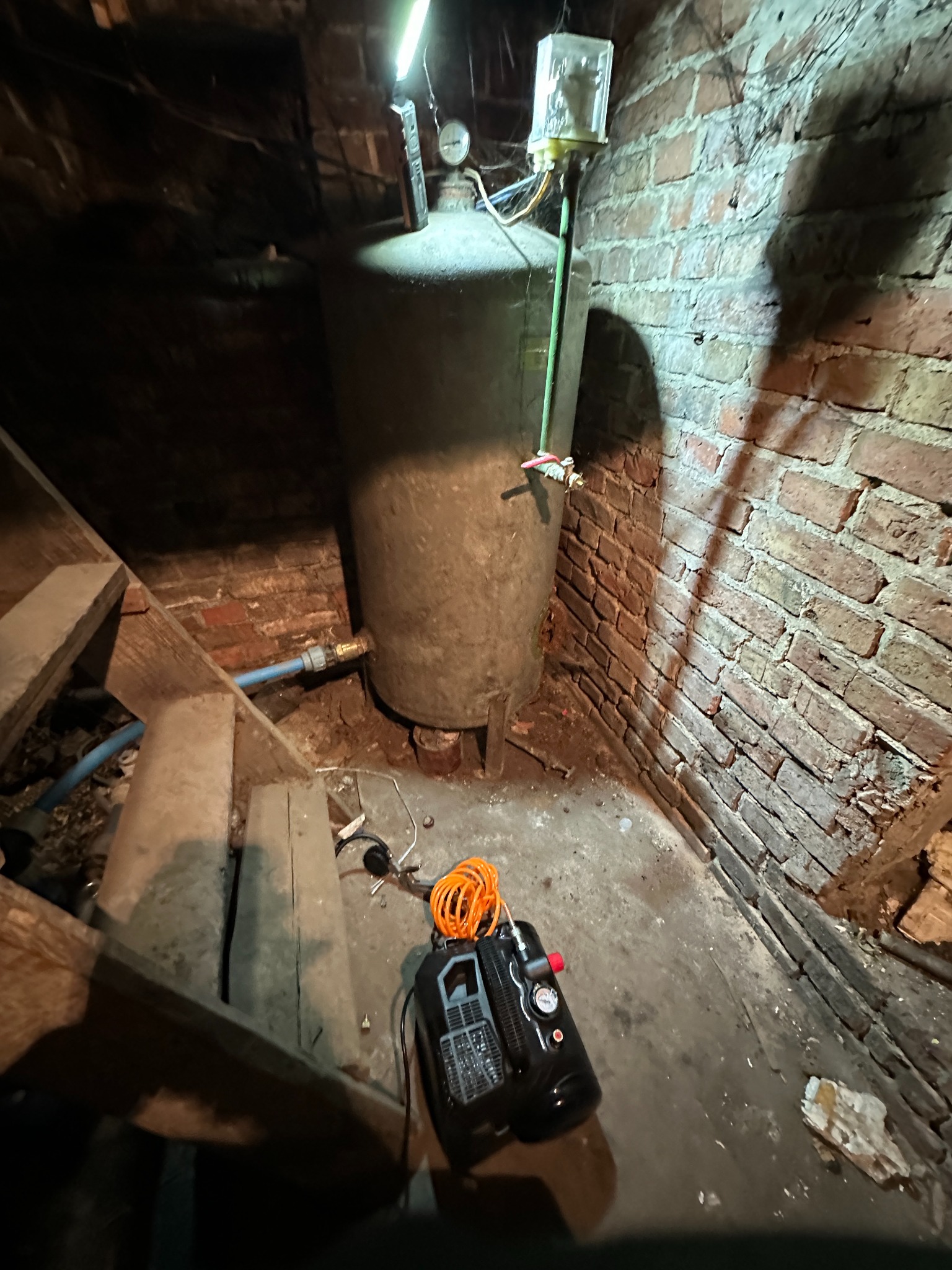 Stary, pionowy zbiornik ciśnieniowy w piwnicy z ceglanymi ścianami, oświetlony lampą warsztatową, obok drewniane schody i kompresor.
