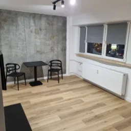 Wnętrze pokoju po remoncie: panele podłogowe w odcieniach beżu i brązu, ściana z surowego betonu, biały grzejnik pod oknem z szarymi roletami, czarny stolik z dwoma krzesłami.