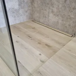 Wykończona łazienka z drewnopodobnymi płytkami na podłodze i wzorzystymi płytkami na ścianie, widoczny odpływ liniowy i fragment szklanej kabiny prysznicowej.