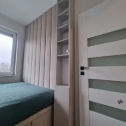 Nowoczesna sypialnia z łóżkiem z zieloną narzutą, panelem ściennym w odcieniach beżu, białą szafką regałową i drzwiami z poziomymi przeszkleniami, widok z boku.