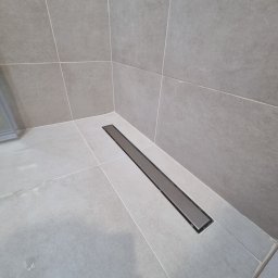 WOJCIECH BIESIADA - Szary odpływ liniowy w narożnej kabinie prysznicowej wyłożonej szarymi płytkami, widoczne delikatne fugi.