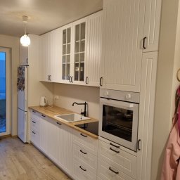 WOJCIECH BIESIADA - Biała, nowoczesna kuchnia z drewnianym blatem, zabudowaną piekarnikiem i szafkami z przeszklonymi frontami, oświetlona wiszącą lampą.